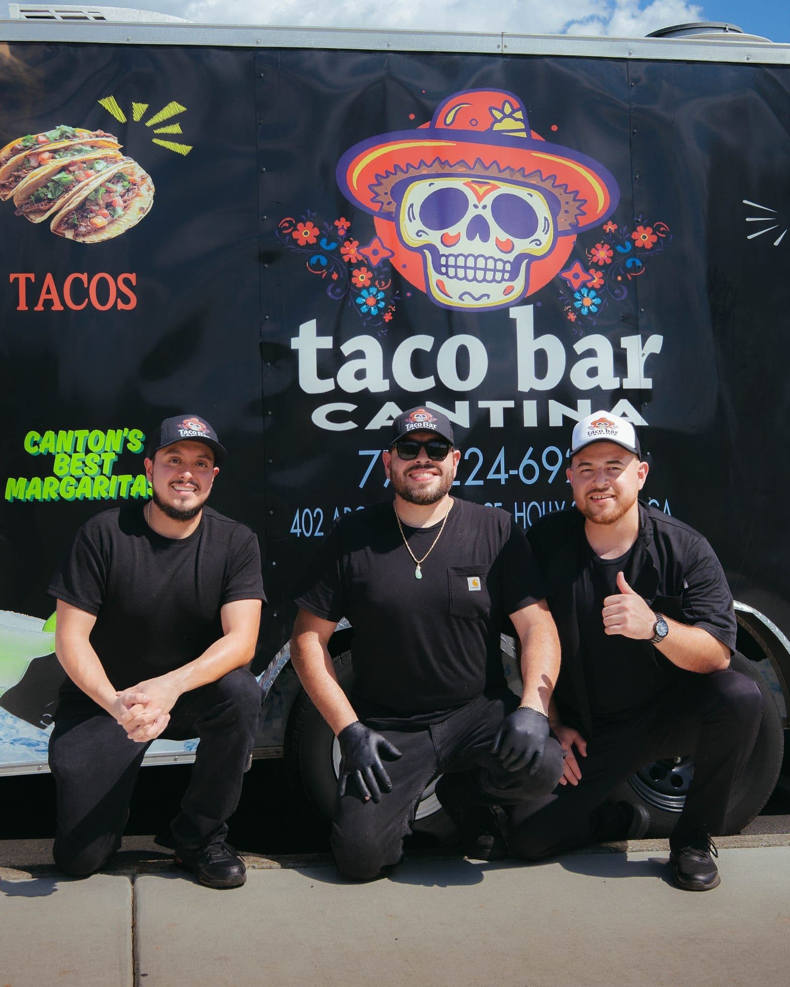 Three Taco Bar Cantina team members crouching in front of the food trailer with the skull logo